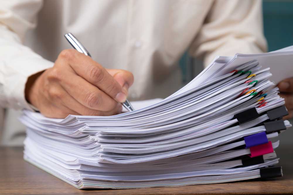 Image of man signing documents on a large stack of papers.
