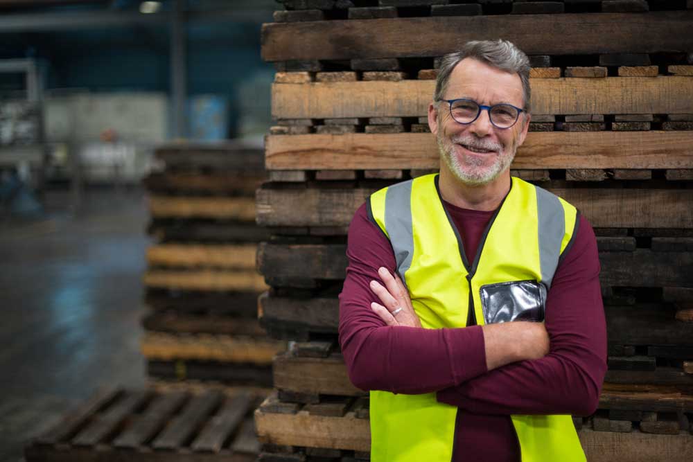 Image of warehouse business owner smiling at the camera, while he leans on wooden palettes. 