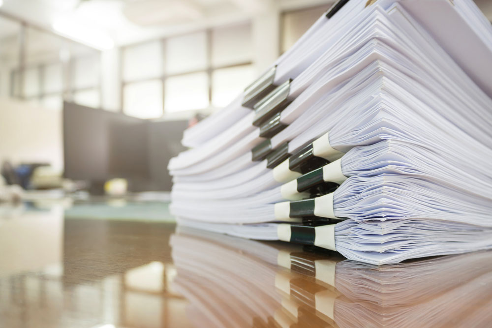 Image of a stack of papers on a desk with computers and a window in the background. 