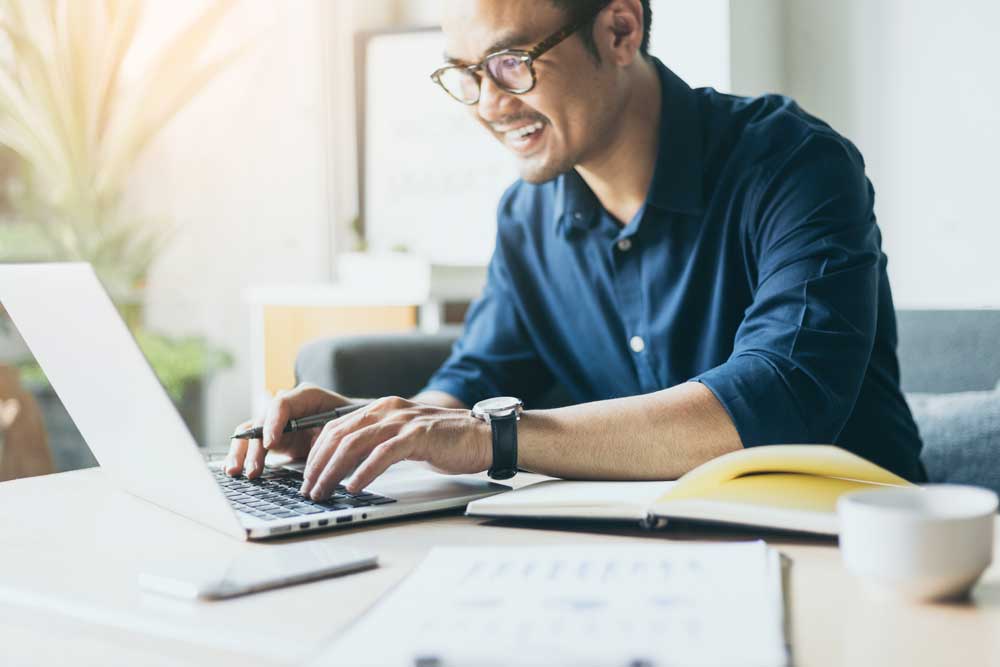 Image of man working on a desk with his computer and a notebook, while he smiles  at the computer. 