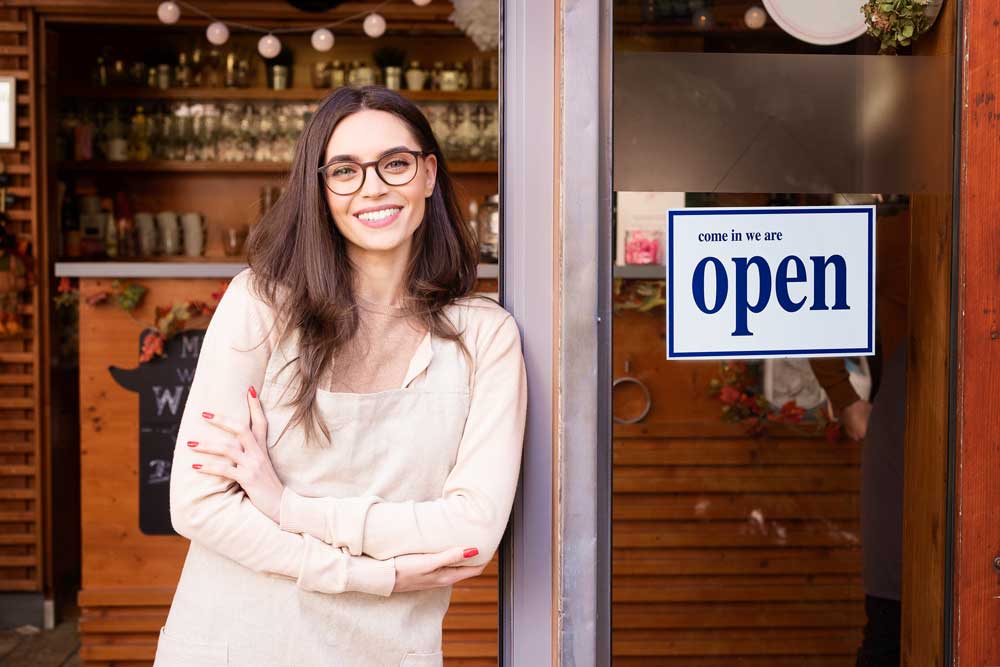 Image of woman business owner smiling at the camera as she leans on her door with a sign that says "come in we are open."