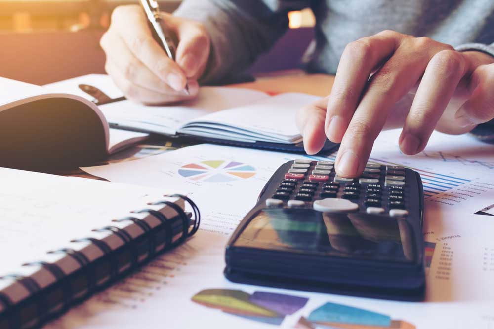 Image of man calculating expenses while he writes them down on a notepad that is stacked on top of graphs.