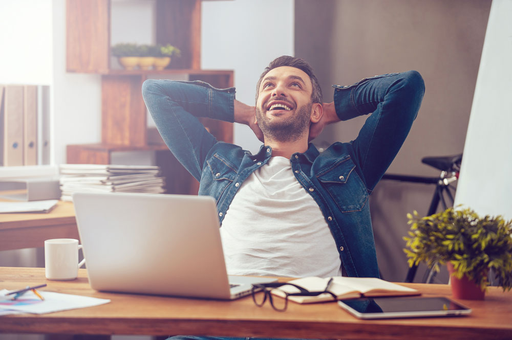 Image of man leaning back on his desk as he smiles up.