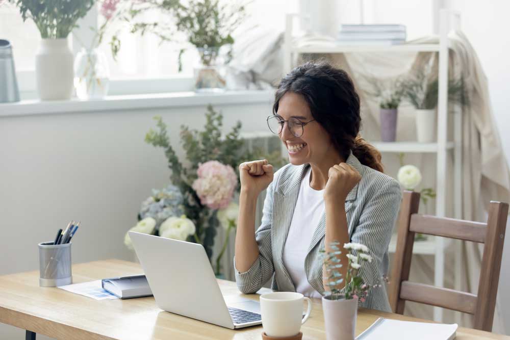 Image of woman smiling as she raises her fists before her. She is also sitting at her desk with her laptop in front of her. 