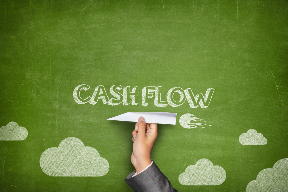 Image of a chalkboard drawing with the word cash flow written on it. Someone holds a paper plane against the chalkboard drawing. 