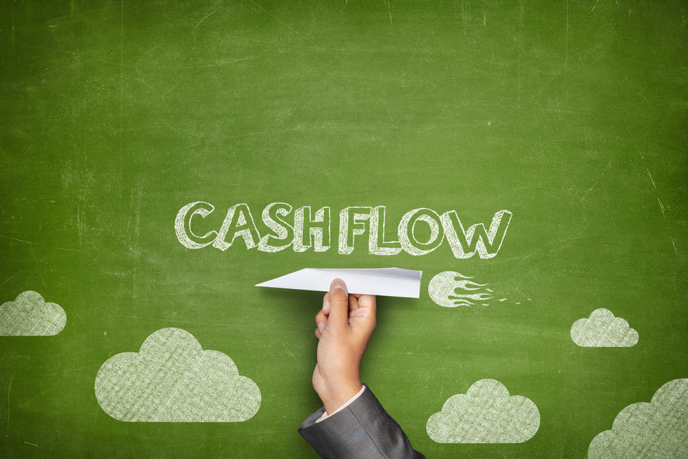 Image of a chalkboard drawing with the word cash flow written on it. Someone holds a paper plane against the chalkboard drawing.