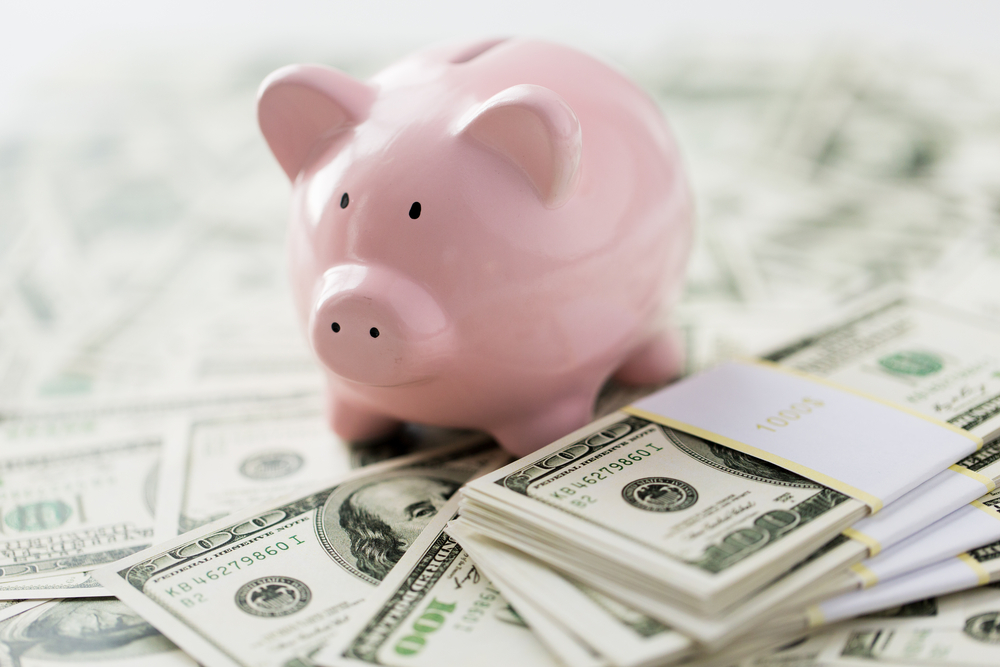 Image of a pile of cash on the floor while there is a ceramic piggy bank on top.