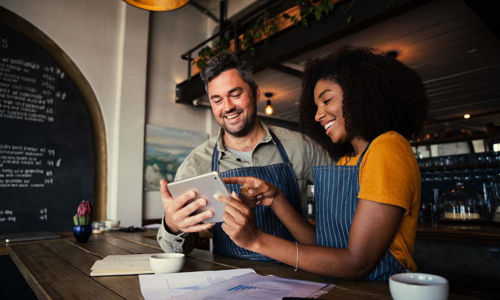 Image of man and woman in a restaurant smiling at an ipad as they both work.