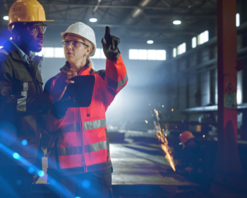 Image of a man and woman in a construction site. The man looks to the horizon while the woman points something out and someone in the background cuts something with sparks flying.