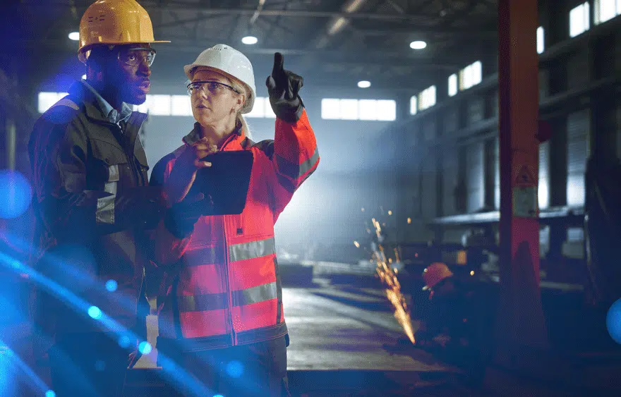 Image of a man and woman in a construction site. The man looks to the horizon while the woman points something out and someone in the background cuts something with sparks flying.