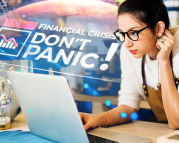 Image of woman leaning over the counter looking at her computer. There is an image in the back that says "Financial Crisis? Don't Panic!"